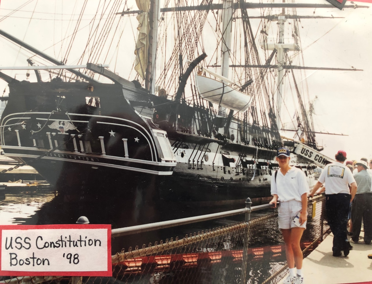 A teenager stands by a fence at a dock in front of a ship from the late 1700s.