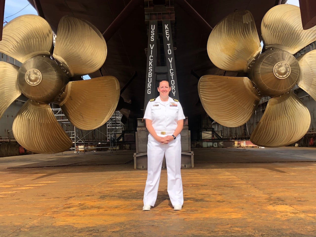 A sailor in dress-white uniform stands in a dry dock in front of a large ship’s propellers.
