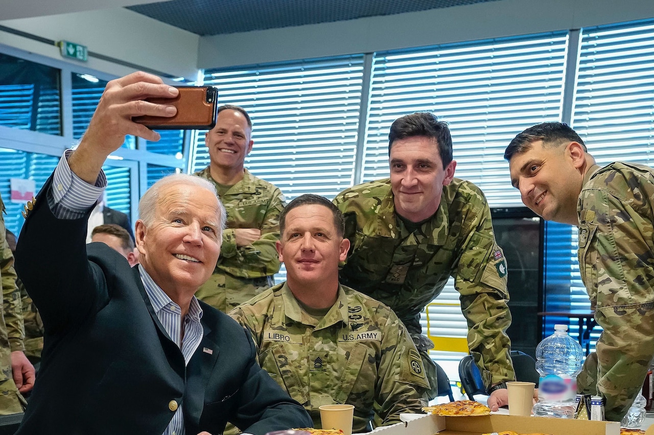 President Joe Biden holds up a phone to snap a selfie with smiling soldiers.
