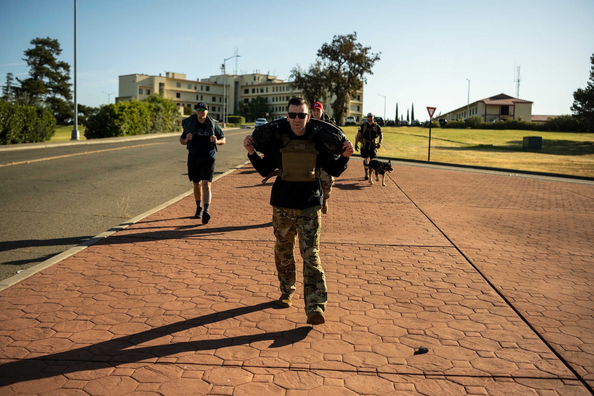 Service members walk along a sidewalk. In the center of the frame a man is seen walking with a large sandbag over his shoulders.