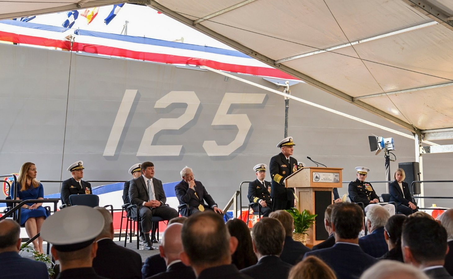 A man speaks from a podium on a stage in front of a Navy ship.