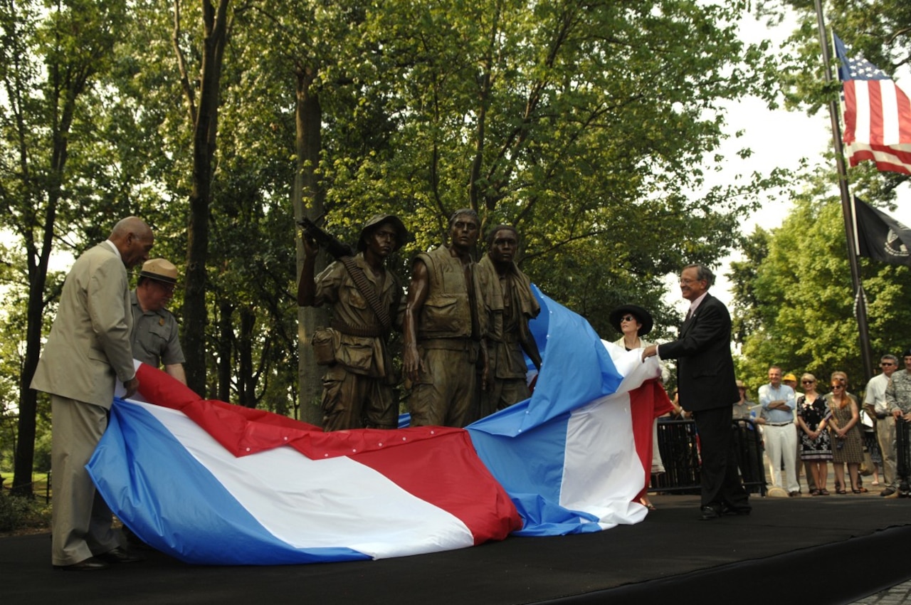 A group of people lower red, white and blue banner to unveil a statue of three men in a park.