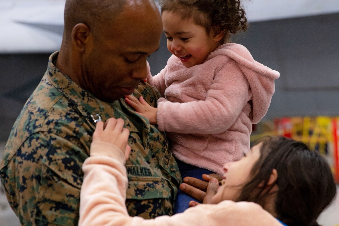 A Marine holds his daughter while another daughter touches a pin on his lapel.