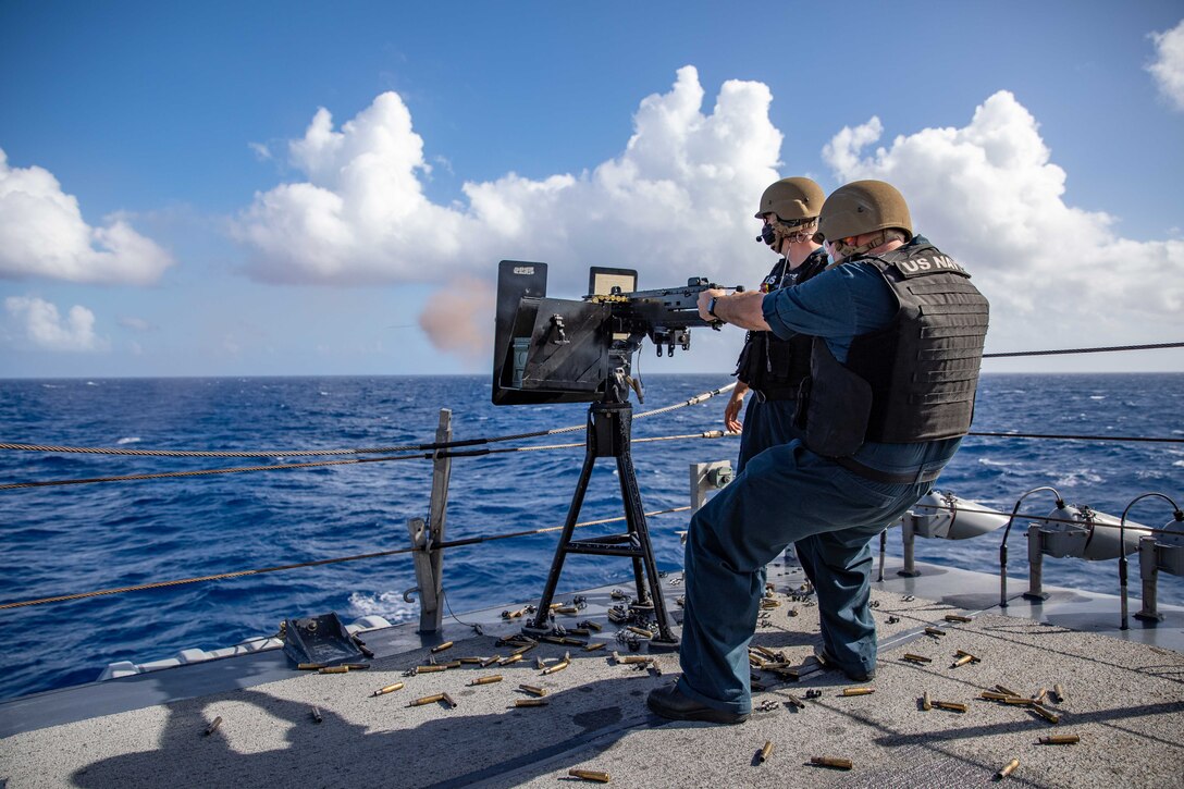 A sailor fires a machine gun while another watches.