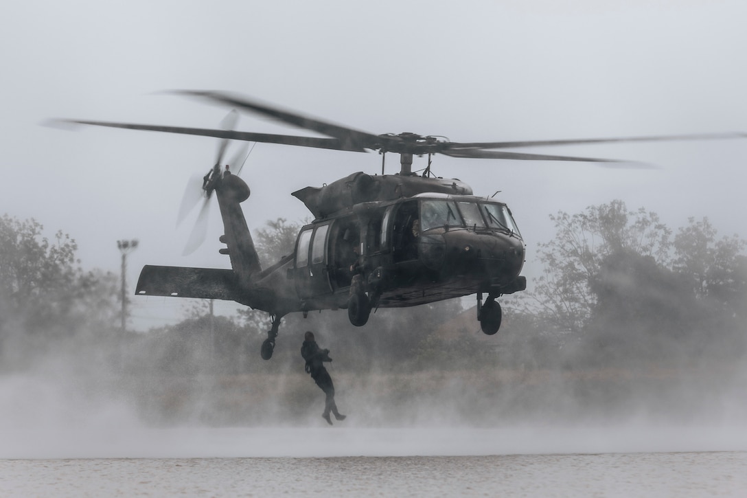 A soldier in a wet suit jumps out of an helicopter hovering over a body of water.