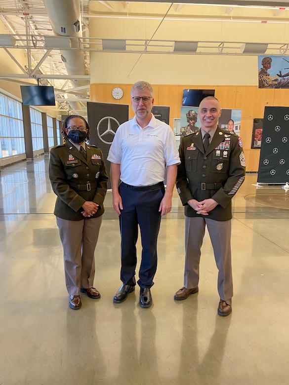 from left to right, woman in U.S. Army uniform stands next to a man in a white shirt who stands next to a man in U.S. Army uniform in the middle of an auto plant.