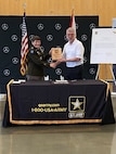 woman in U.S. Army uniform stands holding a plaque with a man in a white shirt behind a table.