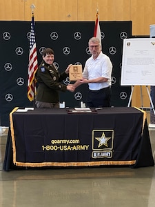 woman in U.S. Army uniform stands holding a plaque with a man in a white shirt behind a table.