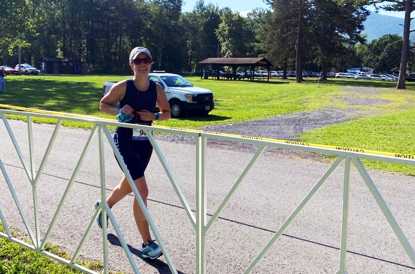 White woman in shorts, tank top and cap runs past a gate in a park.