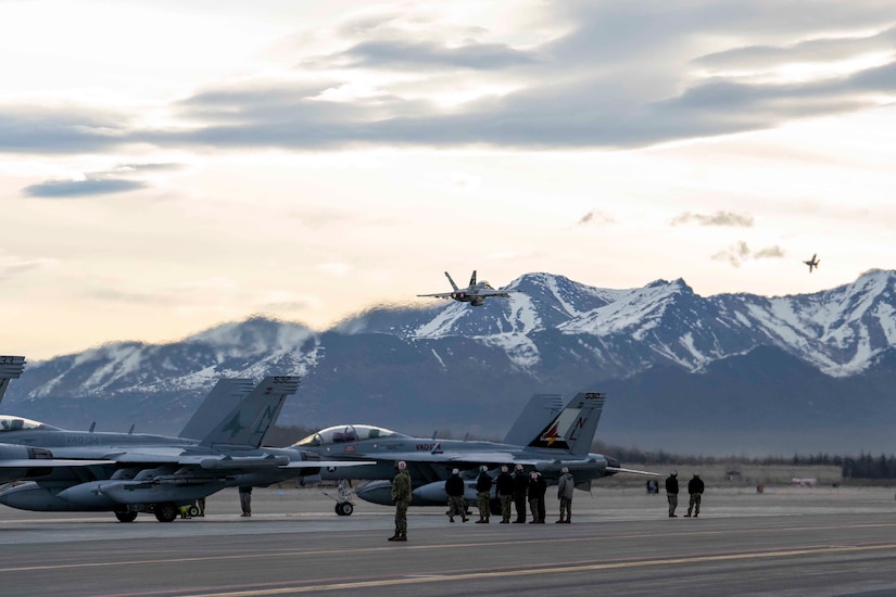 Several aircraft sit on a flightline. Another flies off towards mountains.