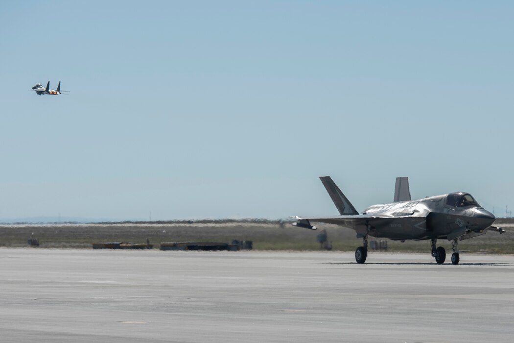 An F-35B Lightning II fighter jet taxis on a flight line and an F-15E Strike Eagle flies in the sky.