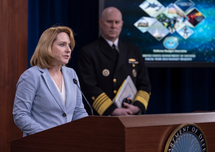 A woman speaks from a lectern.  A man in military uniform looks on from a distance