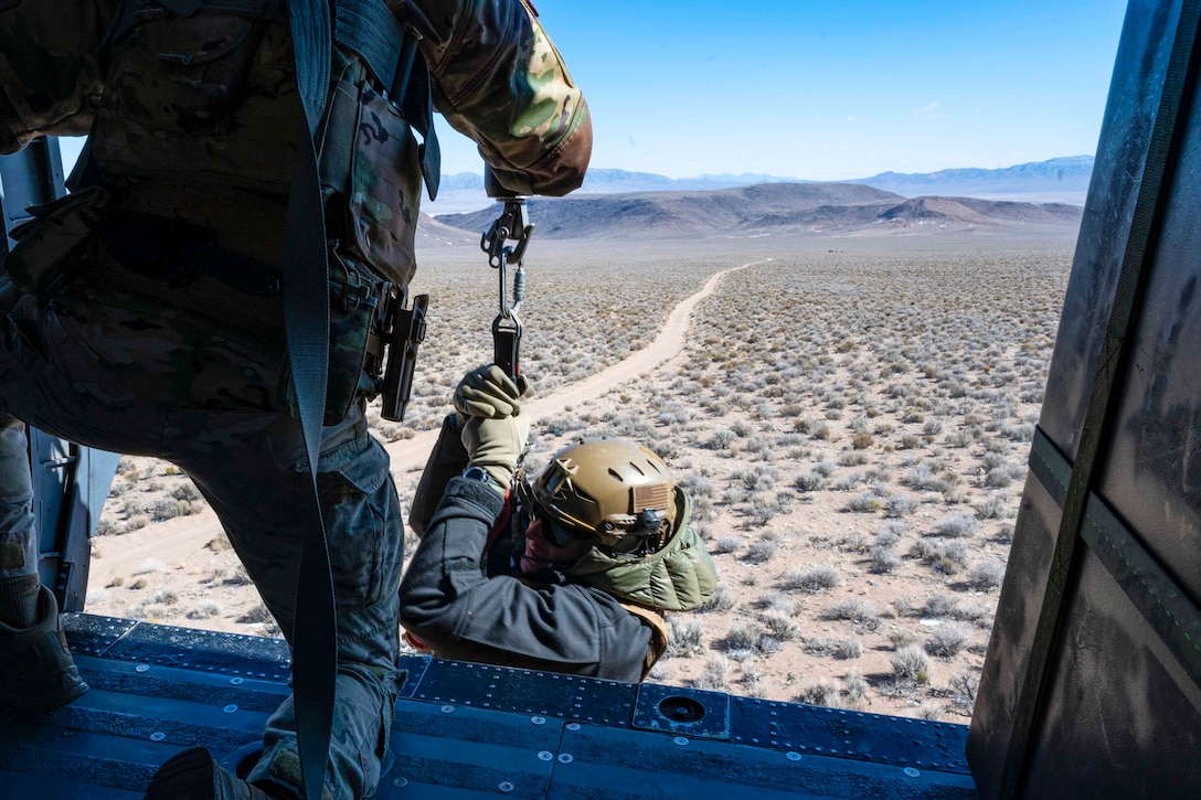 An airman hoists another airman into an airborne helicopter.