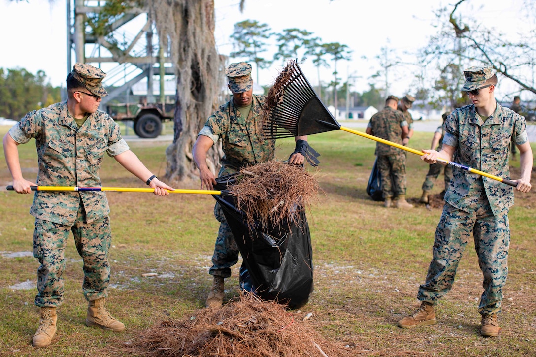 Two Marines rake leaves and debris into a bag that another Marine is holding.