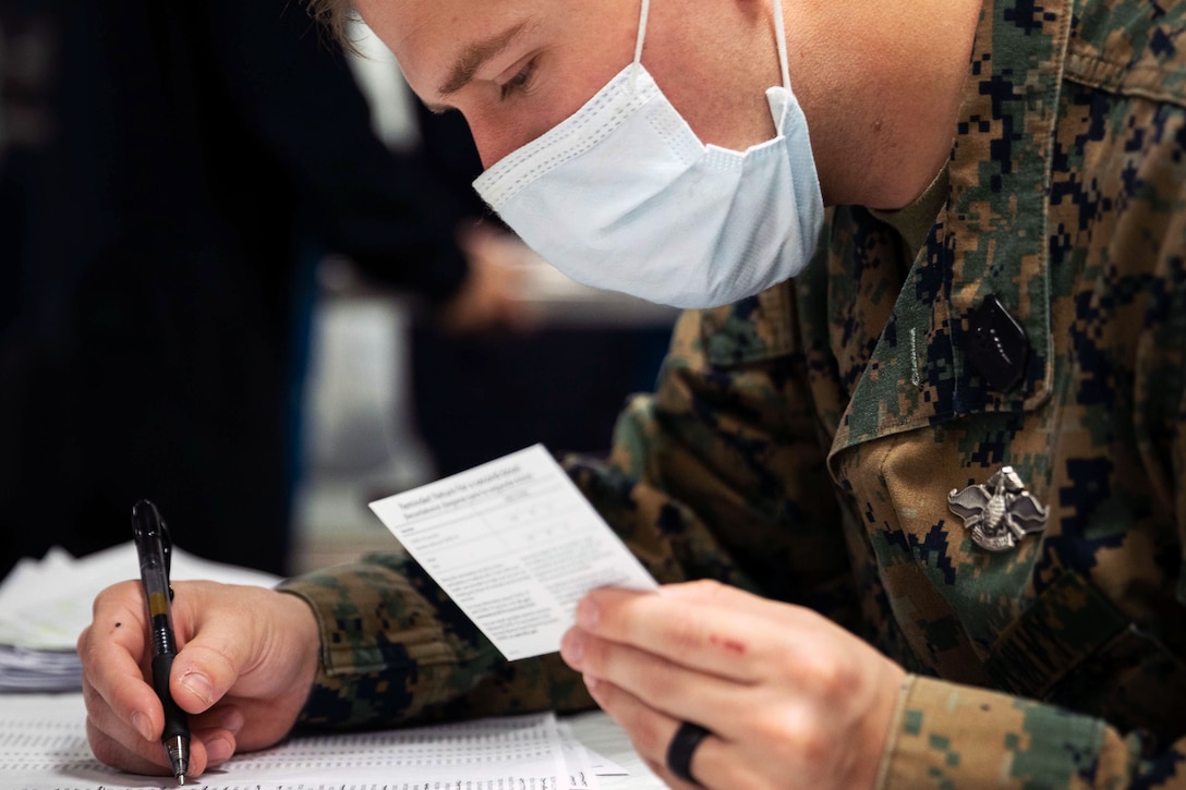 A sailor wearing a face mask holds an ink pen in one hand and a COVID-19 vaccine card in the other.