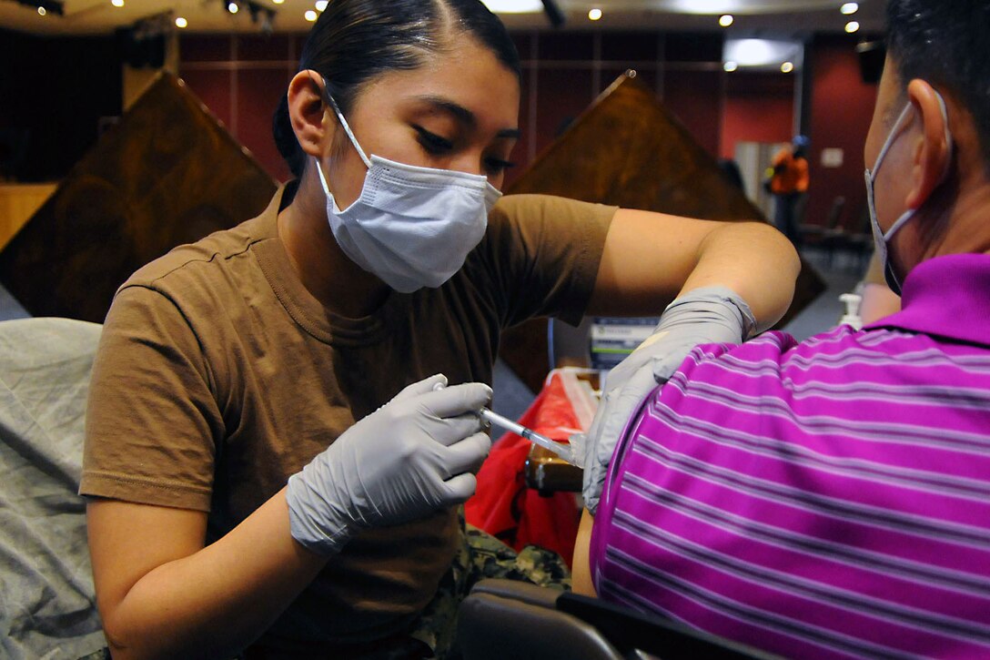 A sailor wearing a face mask and gloves administers a COVID-19 vaccine booster to a local employee.