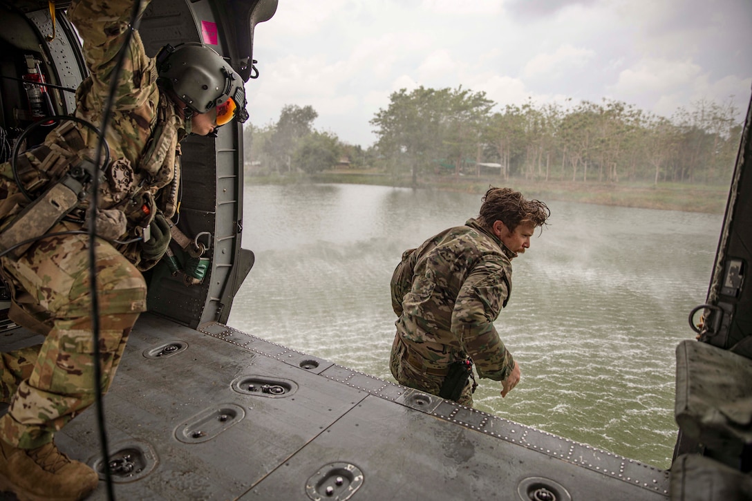 A soldier jumps out of a helicopter hovering over a body of water as a fellow service member watches.