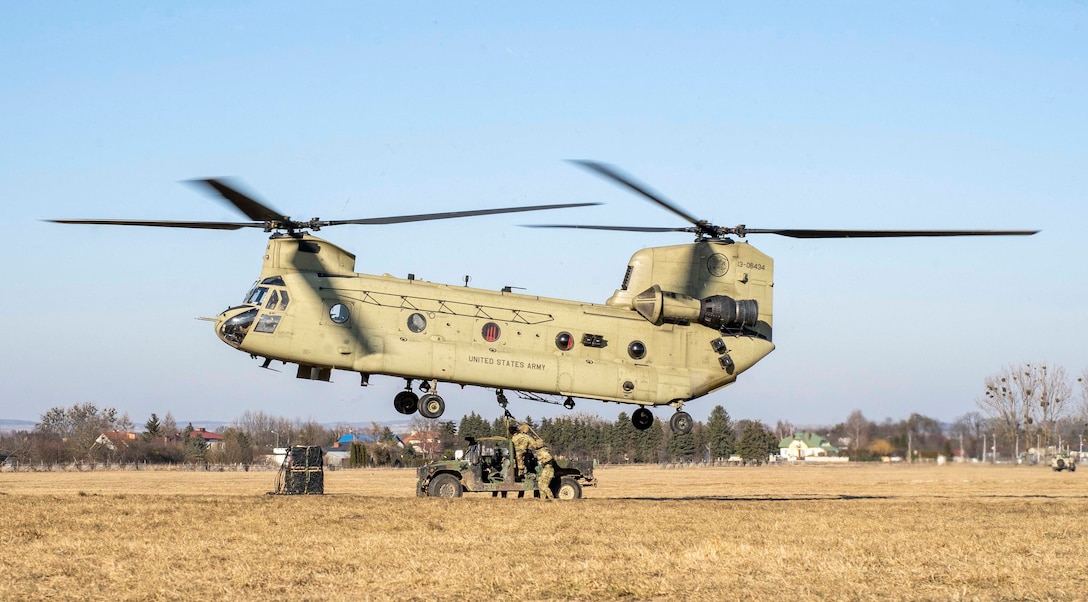 A large helicopter hovers near a military vehicle as soldiers manipulate a line beneath it.