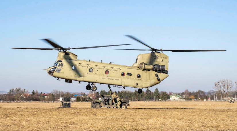 A large helicopter hovers near a military vehicle as soldiers manipulate a line beneath it.