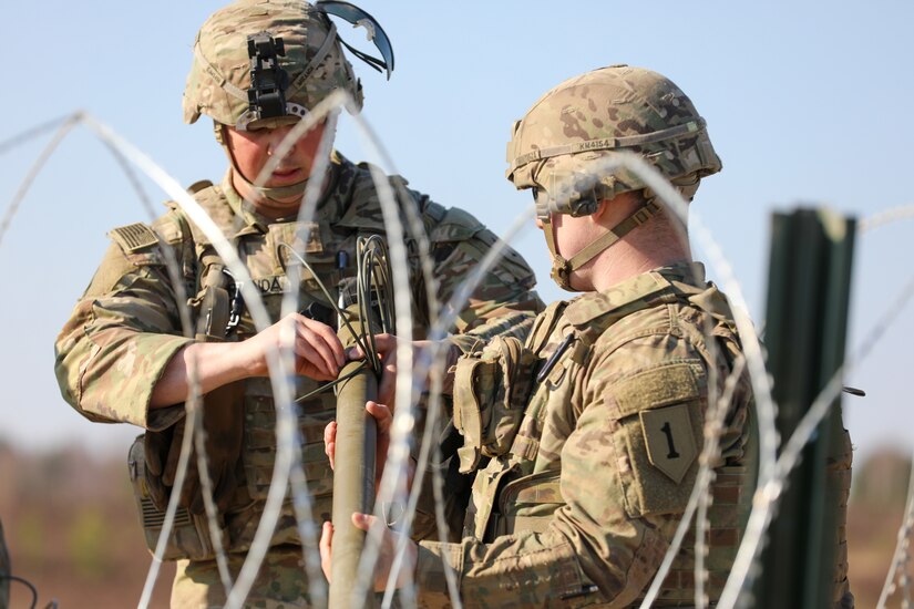 Two soldiers work with what looks like a long, narrow pipe; barbed wire is in the foreground.