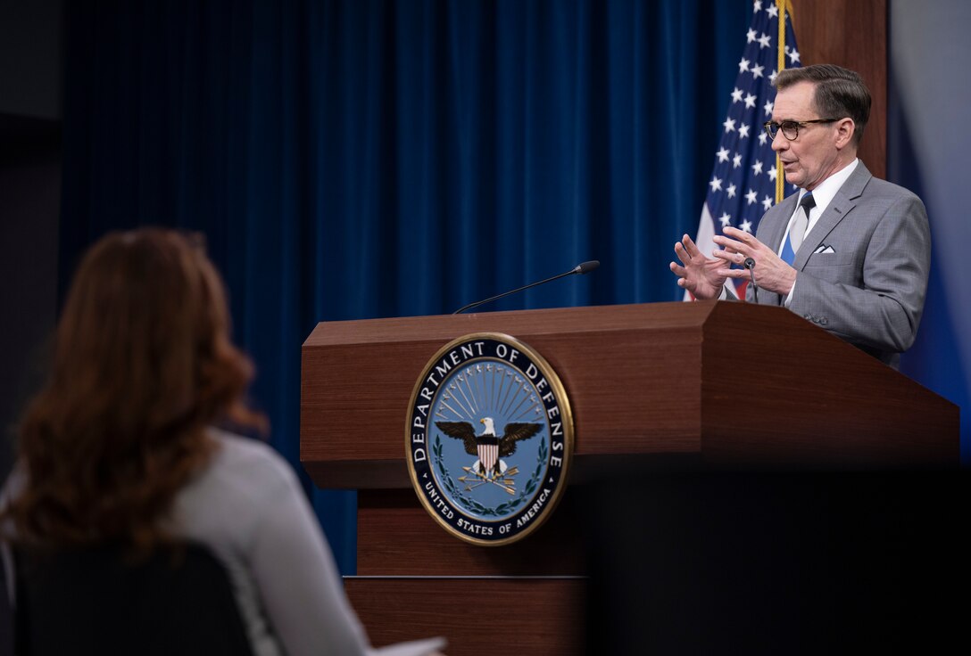 A man stands behind a lectern and addresses seated individuals.