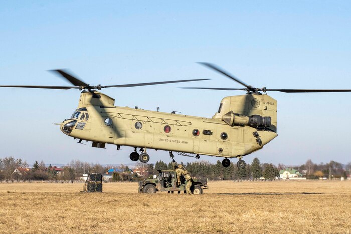 A large helicopter hovers near a military vehicle as soldiers manipulate a line beneath it.