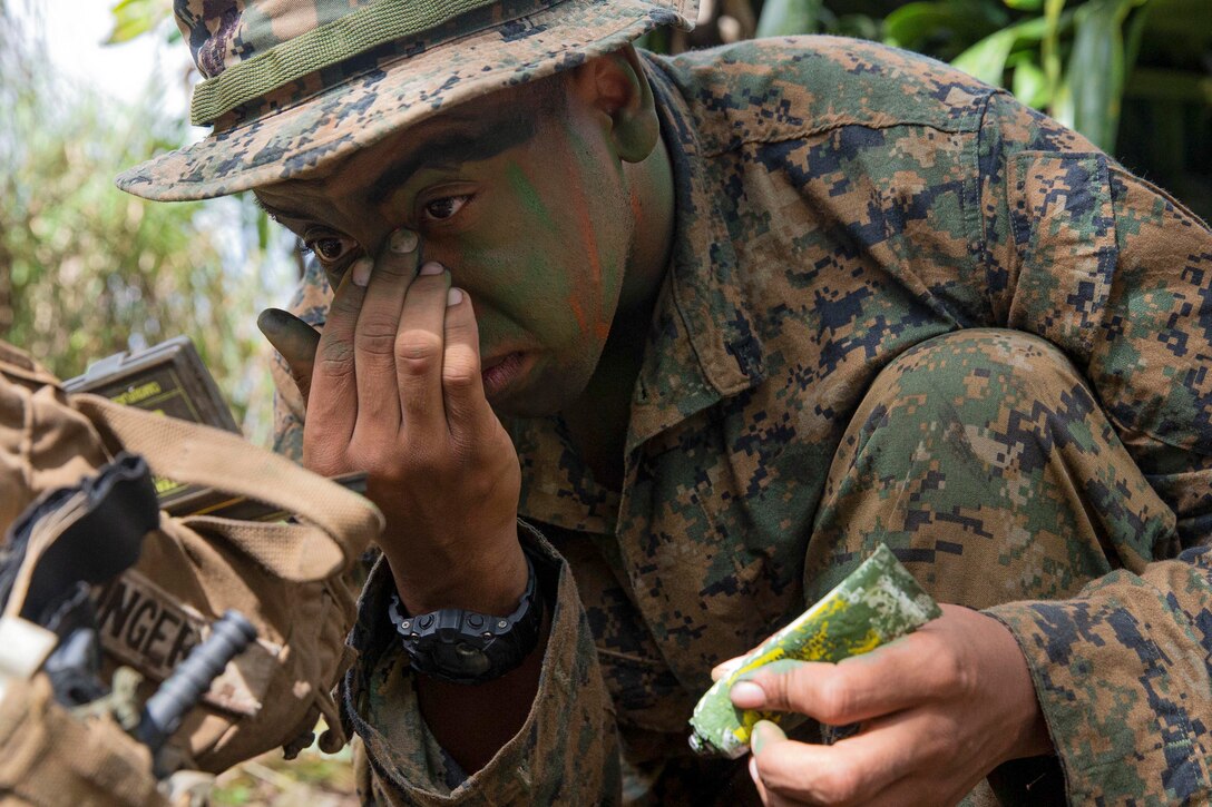 A Marines puts camo paint on his face while kneeling in the jungle.