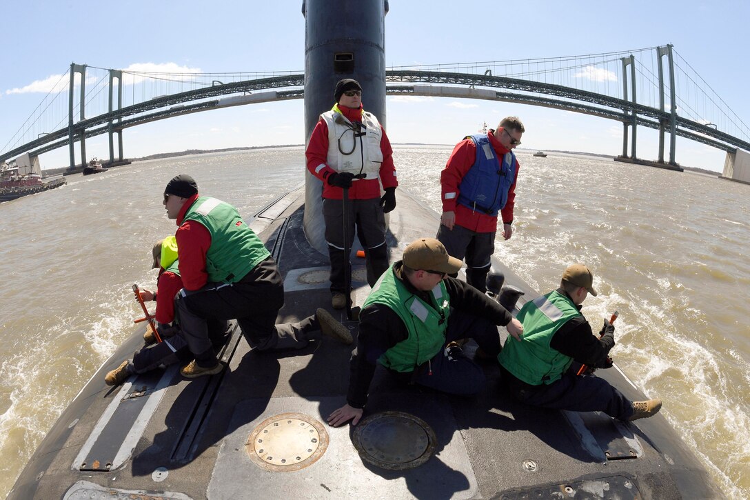 Sailors ride on a submarine near a bridge.