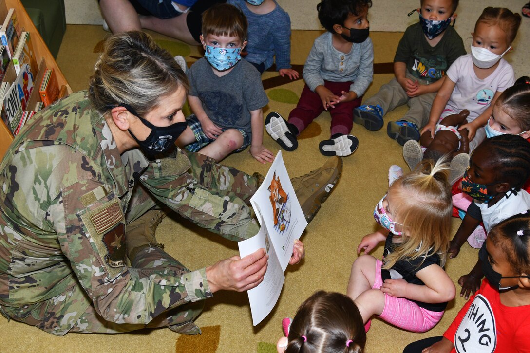 An airman shows a book to children sitting in a semi-circle around her on the floor.