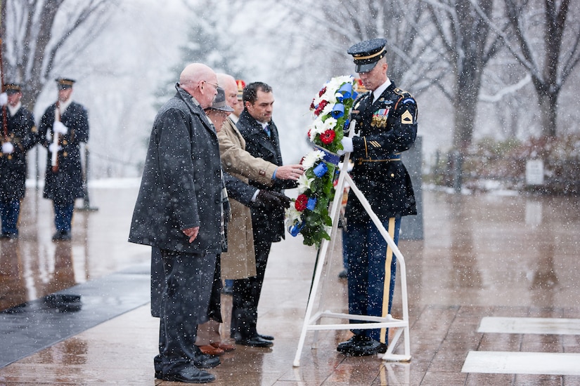 Four men look at a wreath that a soldier has propped onto a stand in a snow-filled plaza.