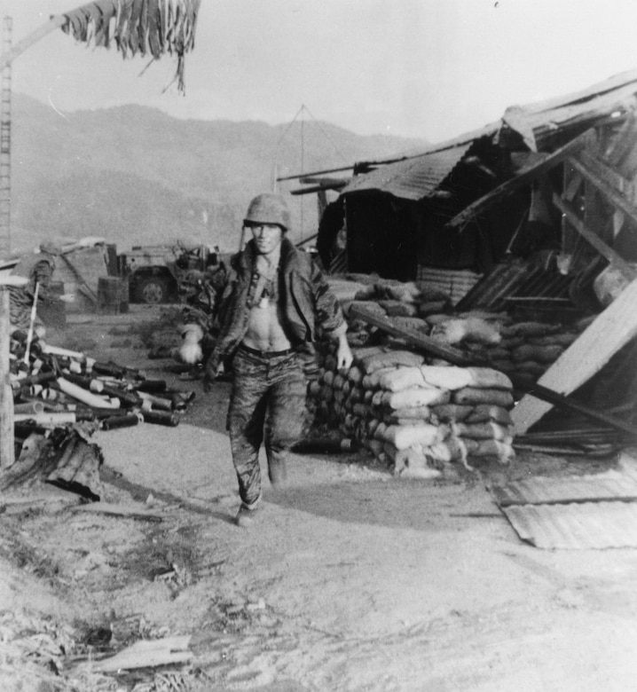 A man in a combat helmet runs past sandbags and a destroyed building.
