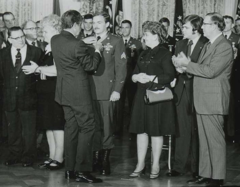 A man puts a medal around another man’s neck while several people watch.