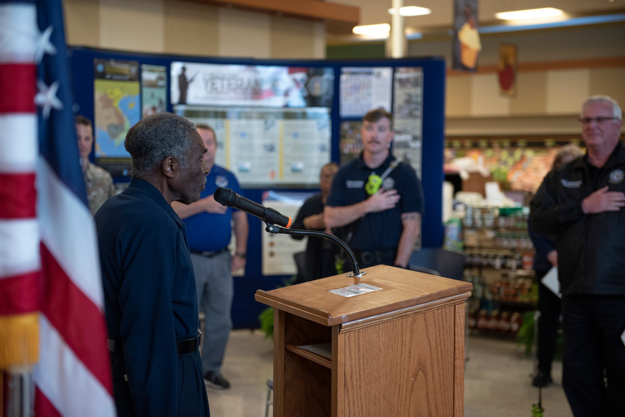 A photo of a man standing at a podium and people standing in front of him with their hands over their heart.