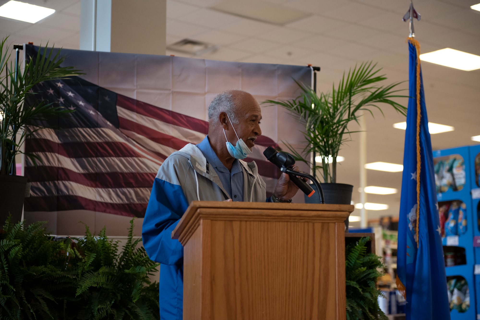 A photo of a man speaking into a microphone in front of a backdrop with an American Flag on it.