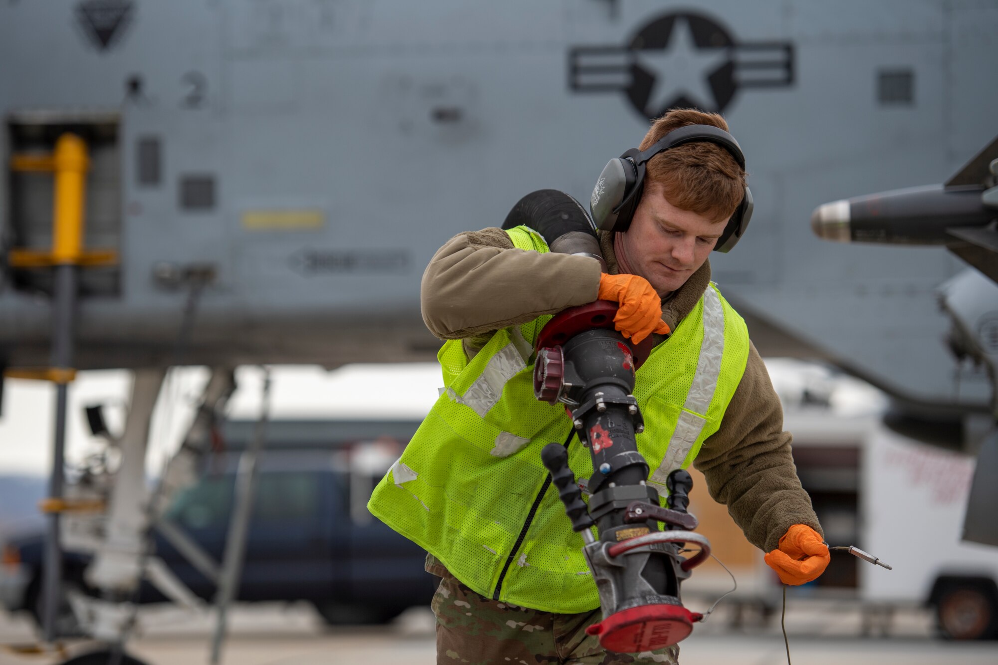 Airman wearing neon green vest pulls a large fuel hose away from an aircraft over his shoulder