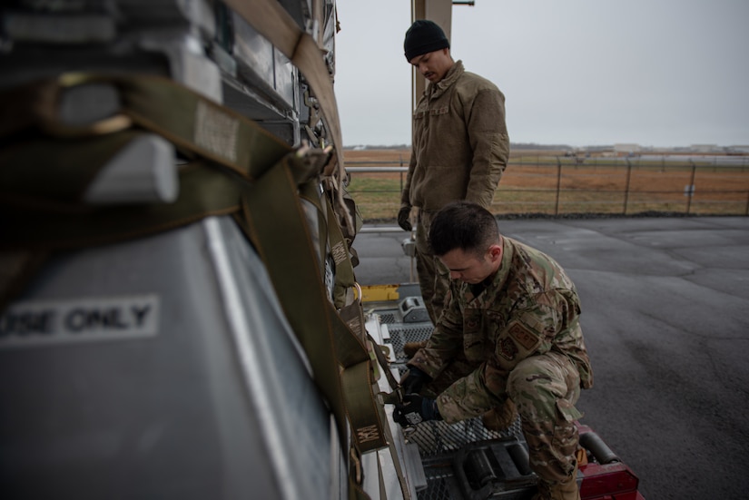 A man in a military uniform adjusts a strap on a cargo pallet. Another man in uniform stands nearby.