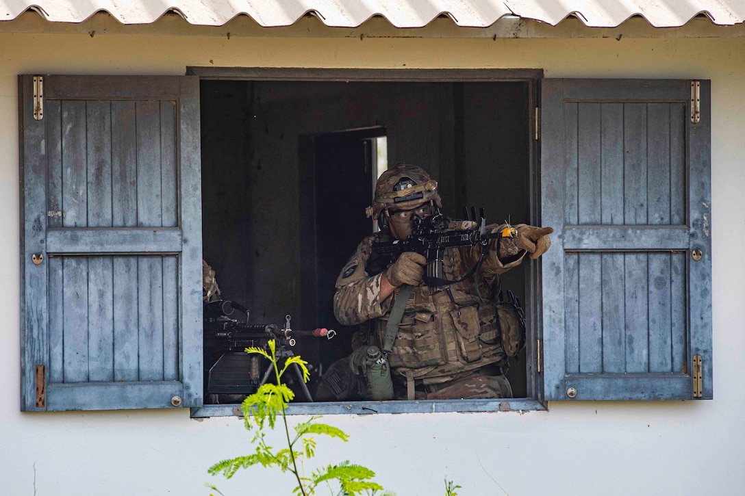 A soldier holding a weapon stands at an open window while pointing out.