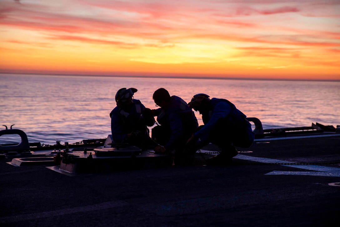 Three sailors shown in silhouette work on the deck of a ship.