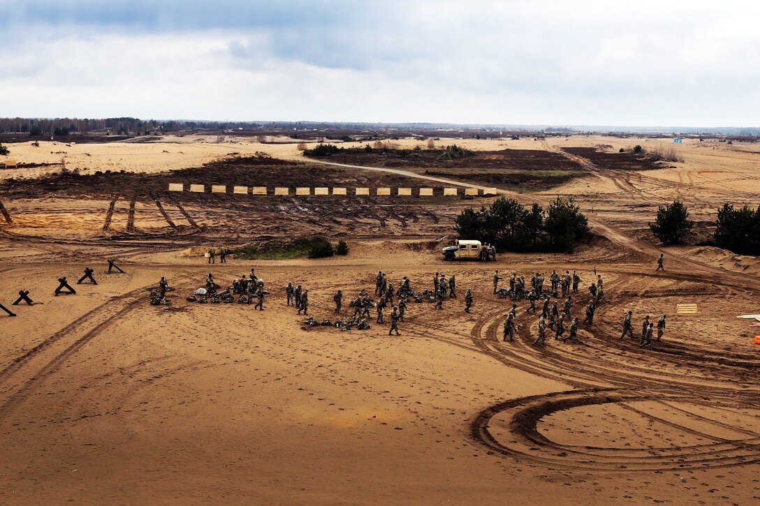 A large group of soldiers walk across an open dirt field that is covered in tire tracks.