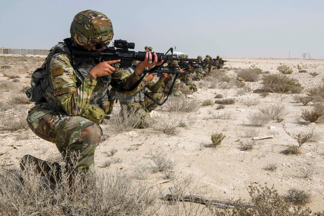 Airmen kneeling in a straight line aim weapons in a desert-like area.
