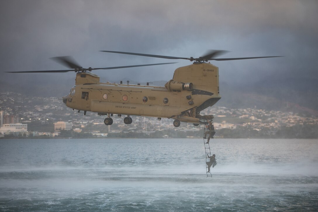 A helicopter hovers over water. Service members descend from the helicopter.
