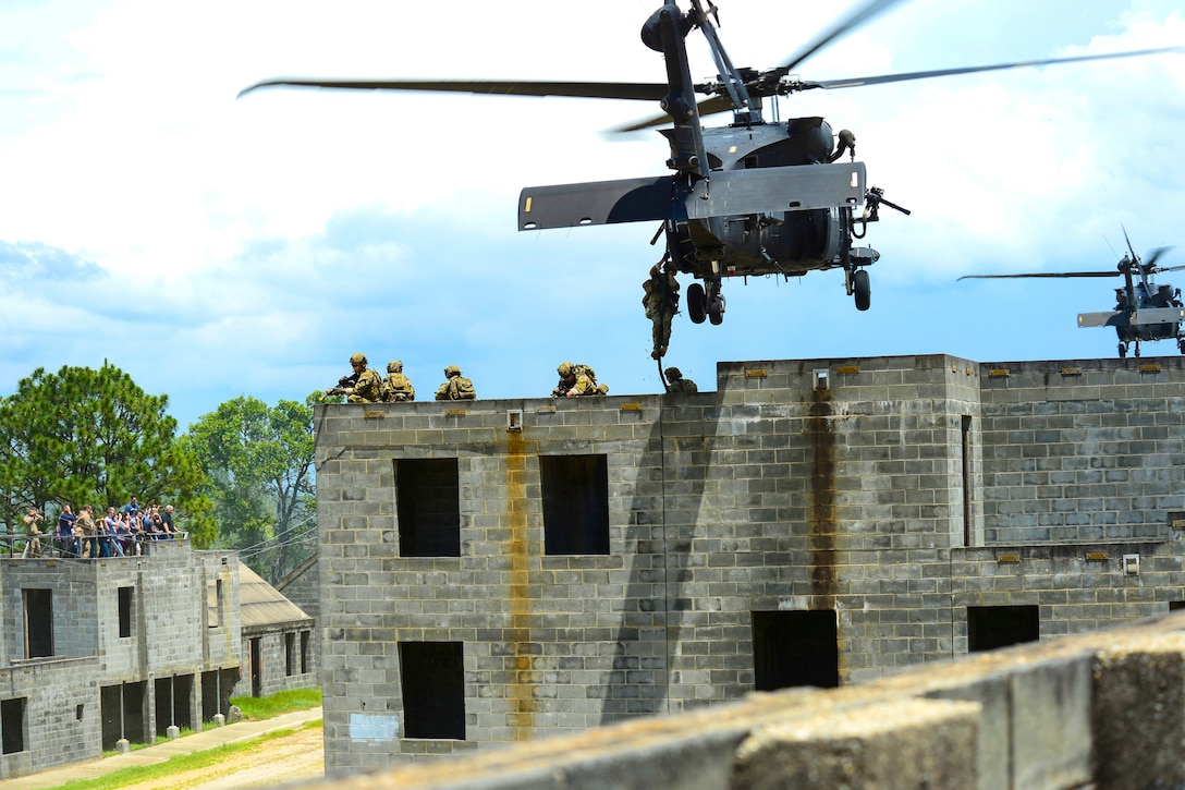 A helicopter hovers over a building. Service members descend from the helicopter.