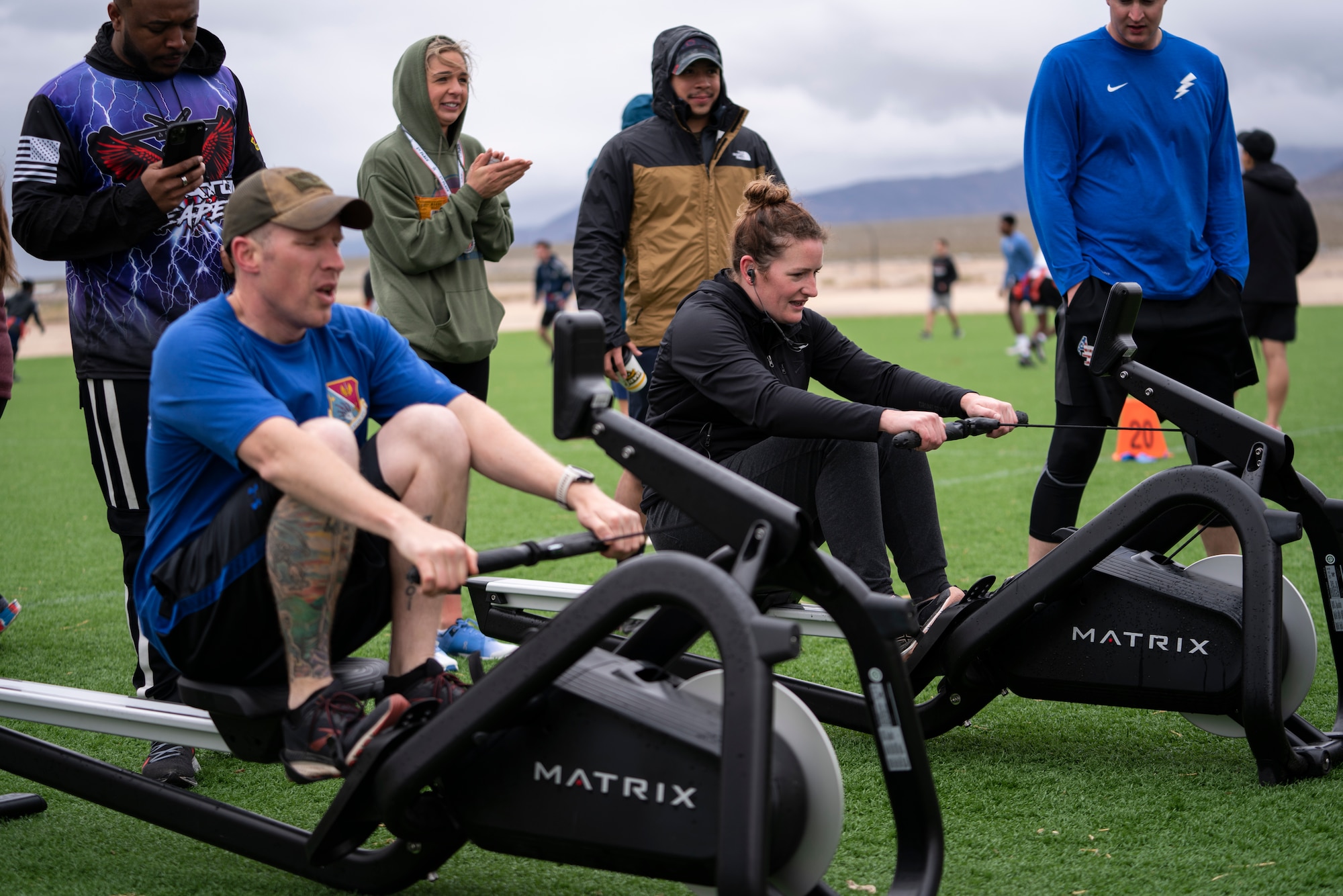 Airmen gather around two people racing on rowing machines.