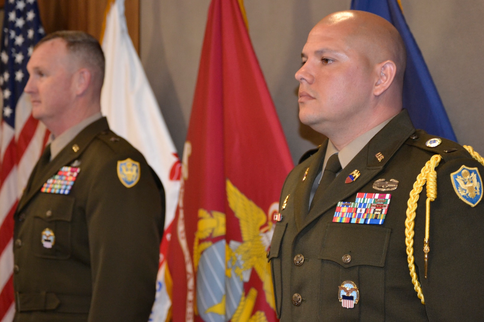 Two men in Army service dress stand at attention in front of a row of flags.