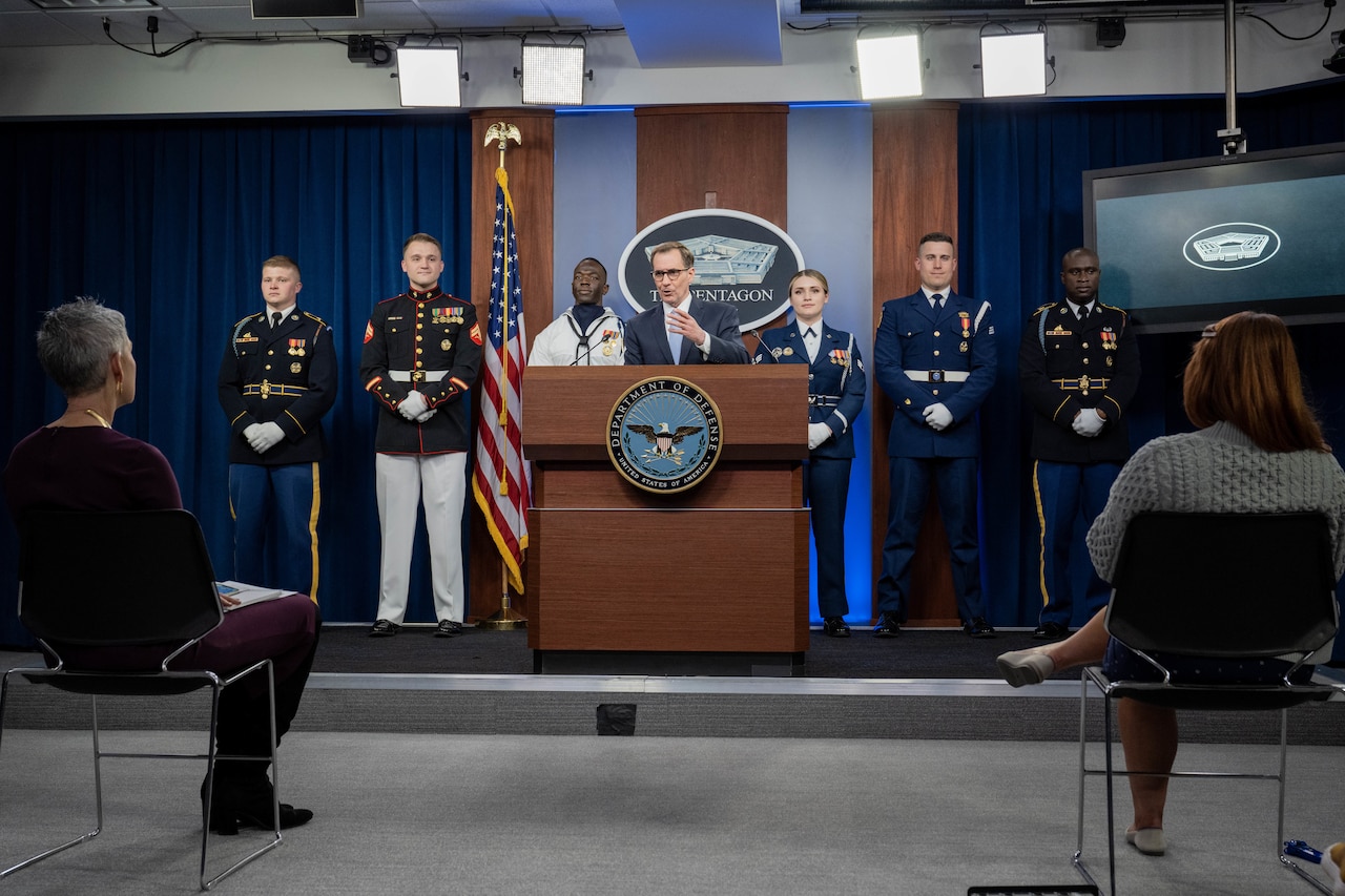 A man stands behind a lectern. Behind him are six military personnel in various uniforms.