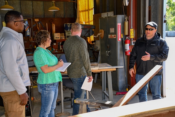 a man speaks to two men and a woman in a carpentry shop