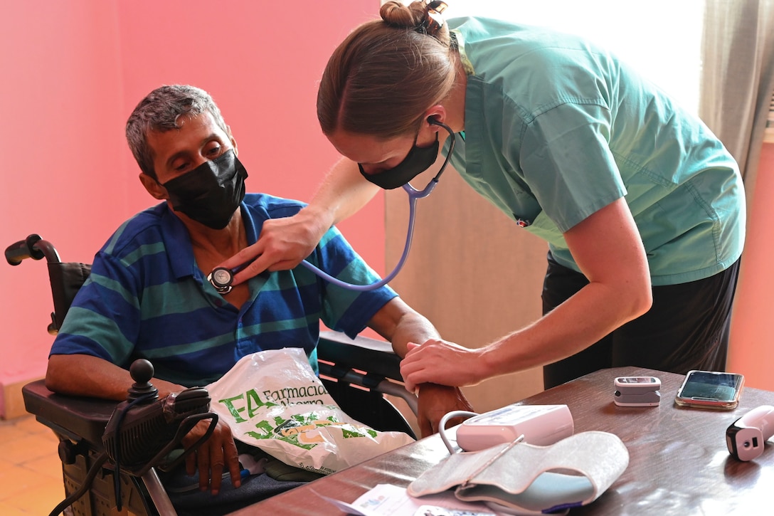 A standing soldier in civilian clothes holds a stethoscope up to a seated man's heart.