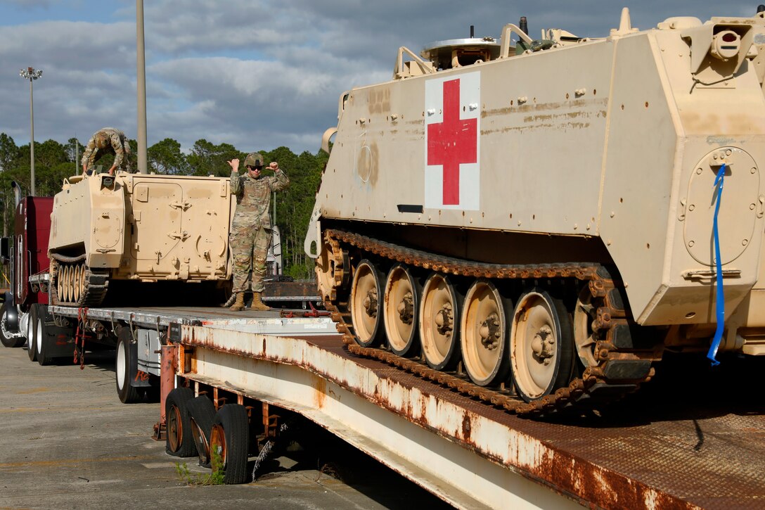 A soldier stands on a flatbed truck and gestures in front of a tank.