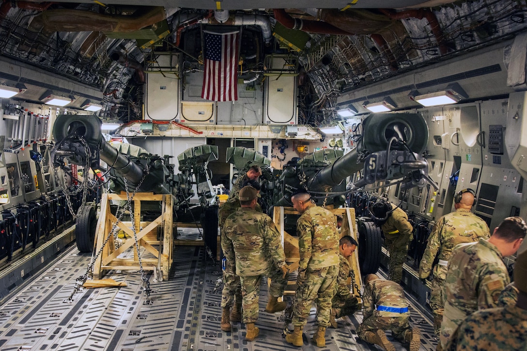 Service members load a large artillery gun into the cargo holding area of an airplane.