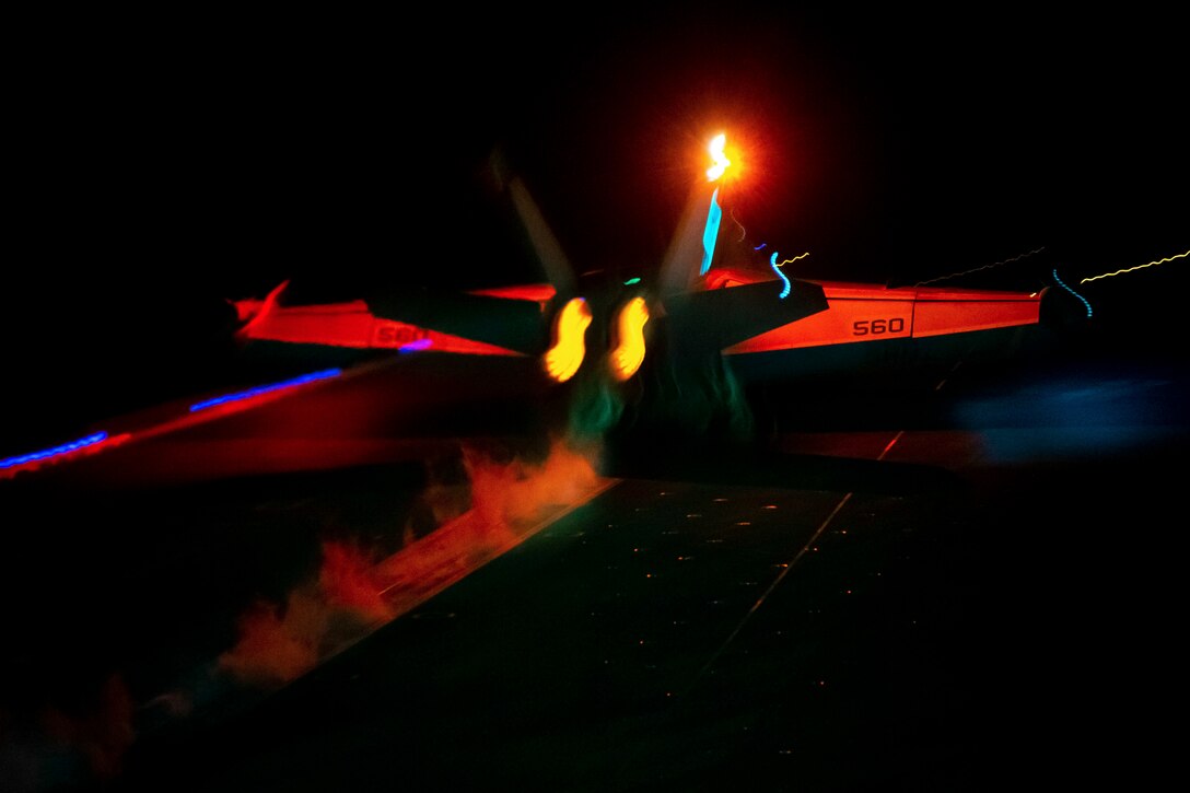 An aircraft takes off from a ship at sea in the dark illuminated by colorful lights.